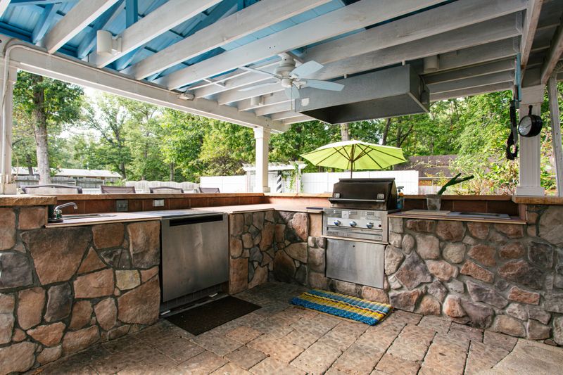 An inviting outdoor kitchen featuring stone countertops, stainless steel appliances, and a ceiling fan, perfect for cooking and dining in a natural setting.