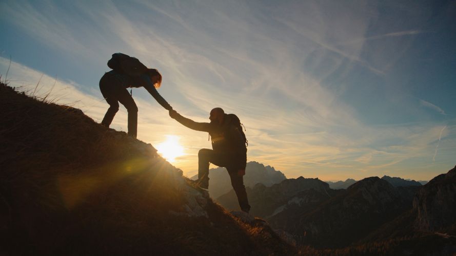 Two hikers helping each other ascend a mountain at sunset.