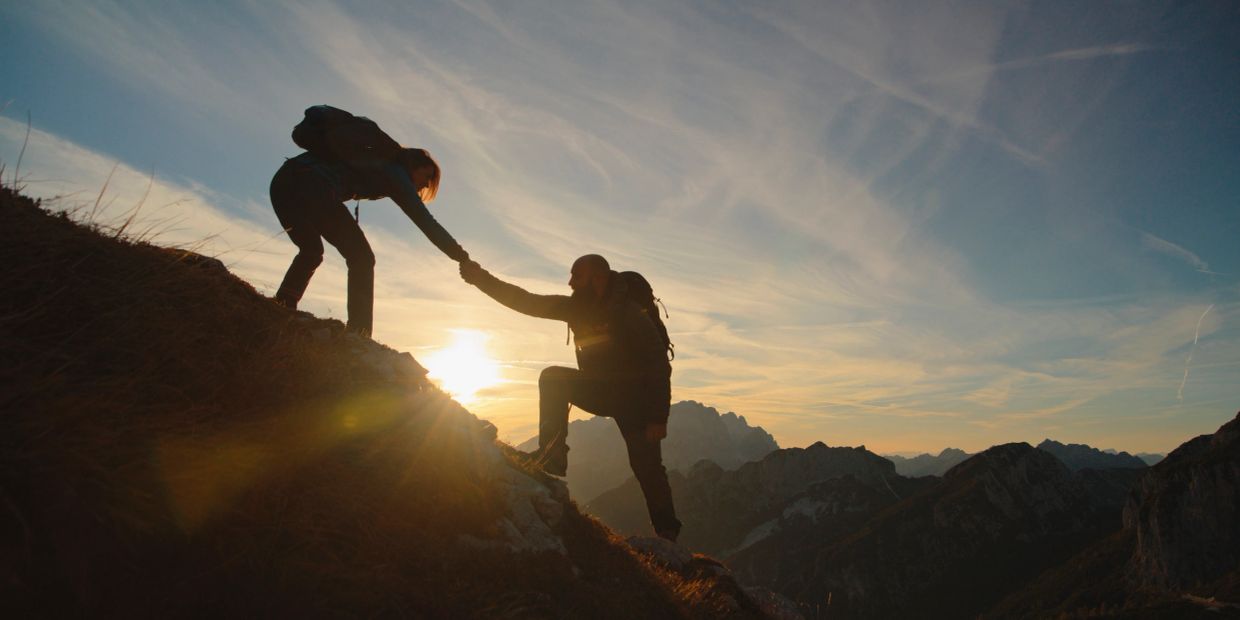 Two hikers helping each other on a mountain slope at sunset.