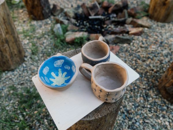Handmade ceramic mugs and a bowl with ocean-themed designs on a wooden stump outdoors.