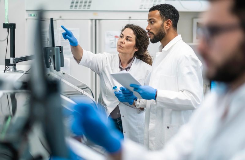 A mid-adult Caucasian woman and a mixed race man, both clad in lab coats, engage intently over research data in a bright laboratory setting.