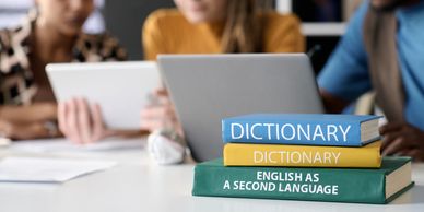 Stack of language and dictionary books with students using tablets in the background.