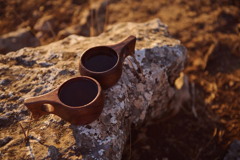 Two handcrafted wooden cups filled with coffee sit on a rocky surface in a serene outdoor landscape, capturing the warm glow of golden hour.