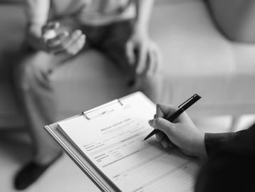 A person fills out a medical history form while another person sits on a couch.