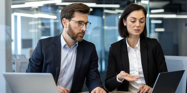 Two professionals in business attire discussing work on laptops in an office.