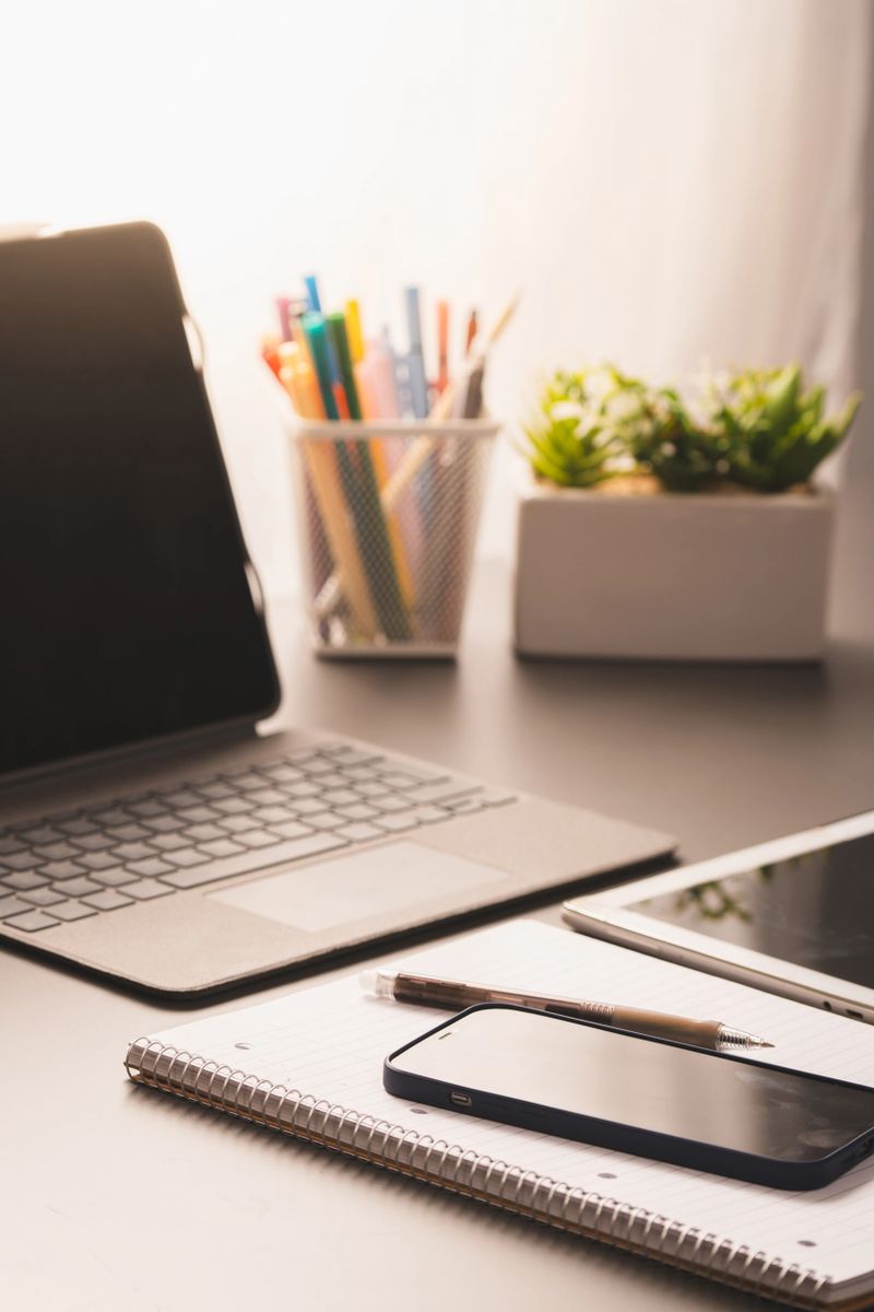 A modern home office desk featuring a laptop, notebook, smartphone, pen, and office supplies. Perfect setup for remote work or studying - Bright and organized workspace with natural light.