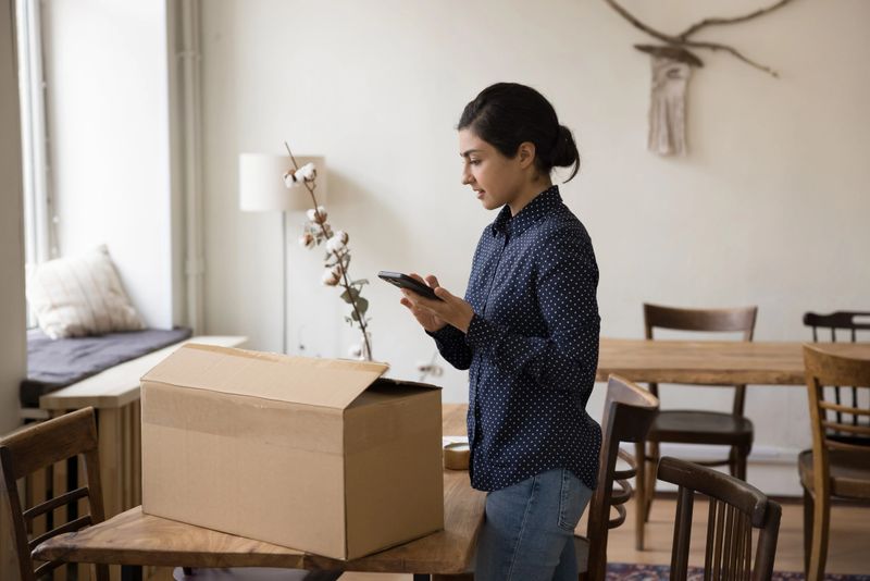 Young Indian woman prepare cardboard box for shipment, hold cellphone, mobile application for sending parcel all around world using easy, comfort, trusted transporting courier company on-line services