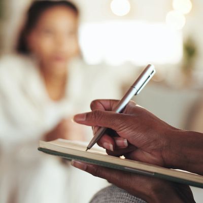 Person writing notes with a pen while someone talks in the background.