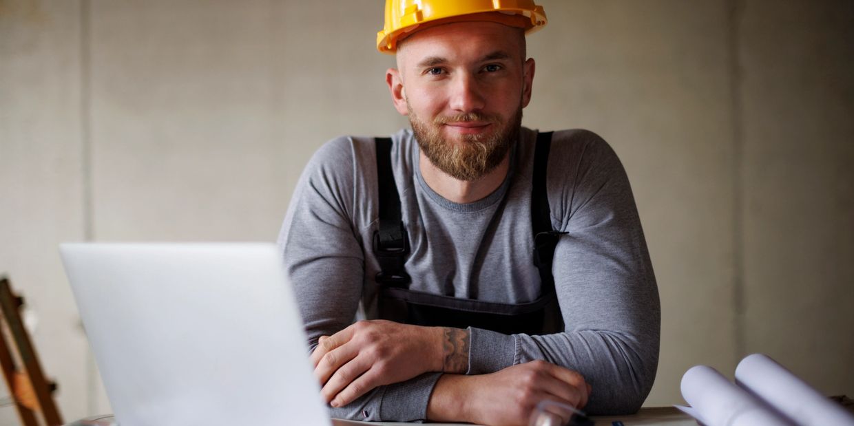Man in a yellow hard hat working at a desk with blueprints and a laptop.