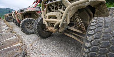 A row of muddy off-road vehicles parked on gravel near a stone wall.