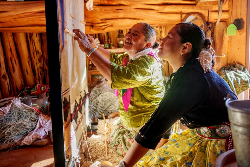 A Navajo Grandmother Teaching Her Teenage Granddaughter How To Weave a traditional blanket On A Loom in a Navajo Hogan in Monument Valley, UT wearing traditional clothing