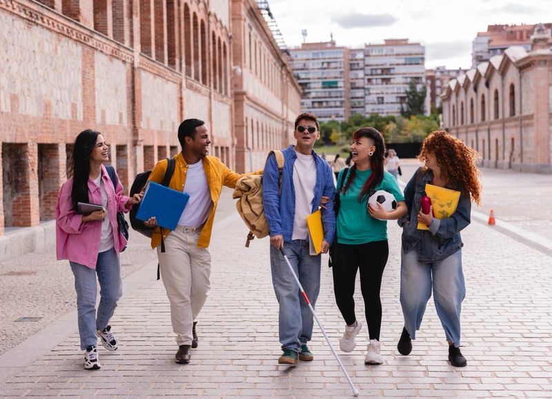 group of diverse young adult students, including one with visual impairment, walking and laughing on a university campus.