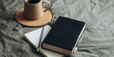 Several items sit on top of a covered bed - a cup of tea, a notebook and a pencil, and a closed book 