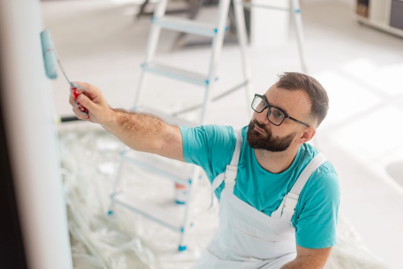 A bearded man in glasses and a blue shirt is painting a white wall with a roller while kneeling, with a ladder and painting supplies visible in the background.