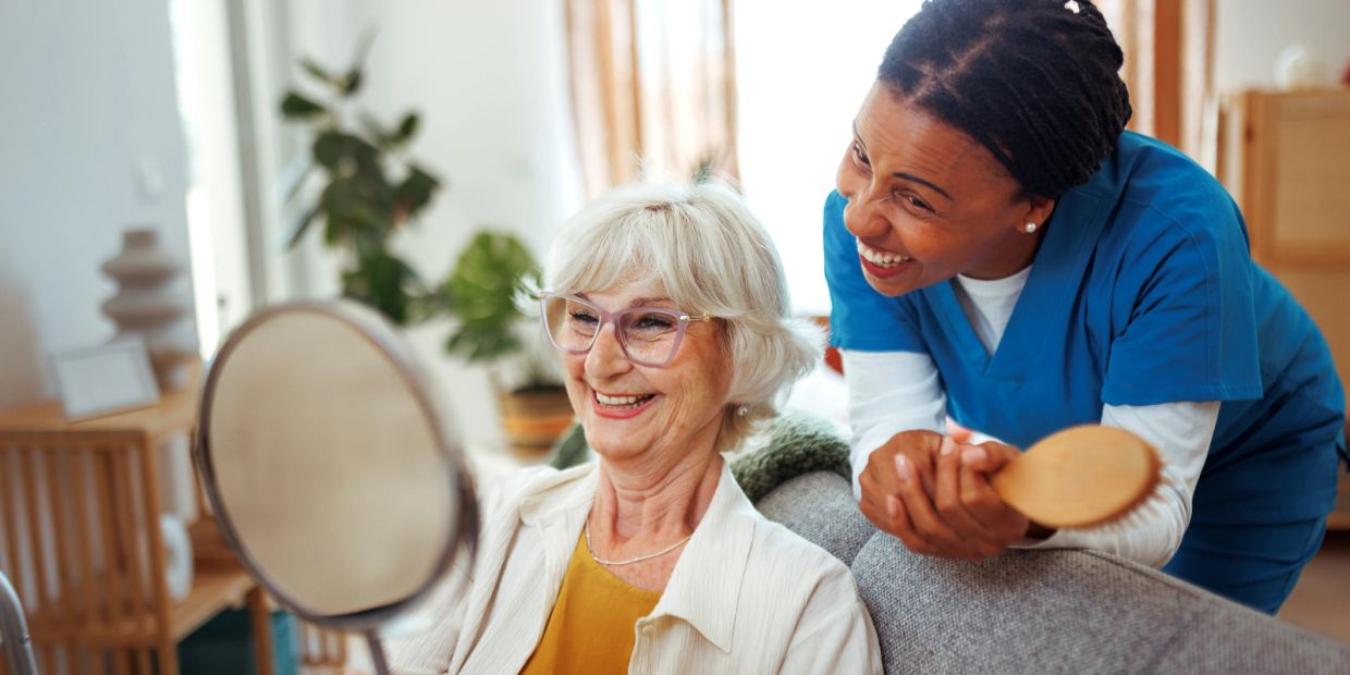 Elderly woman smiling while holding a mirror with a nurse beside her.