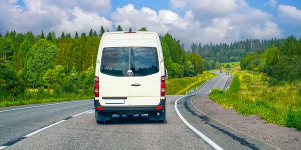 White van driving on a scenic highway surrounded by green trees and blue sky.