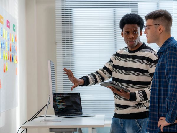 Two developers discussing code in a modern office with sticky notes and computer screens.