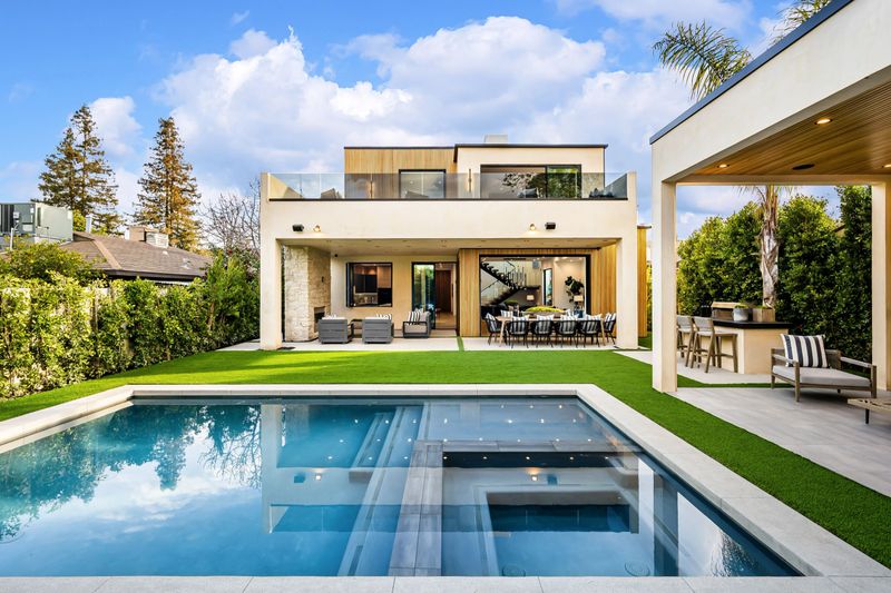 Modern luxury house with a swimming pool, outdoor seating area, and lush greenery under a bright blue sky.