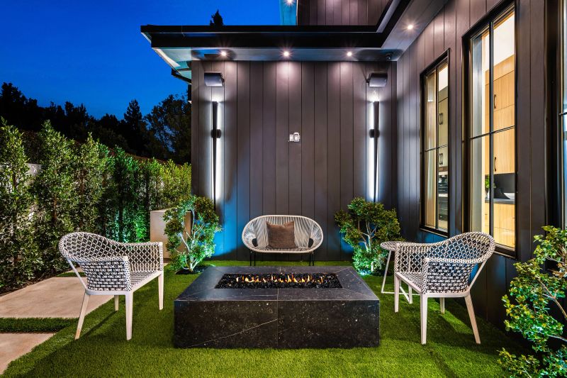 Modern outdoor patio with stylish furniture and a fire pit, surrounded by greenery and illuminated at night.