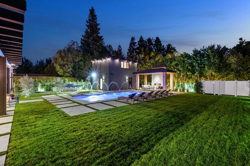 A modern backyard with a swimming pool, lounge chairs, and a patio area illuminated at dusk.
