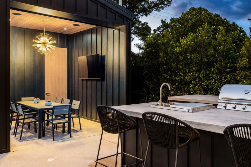 Modern outdoor kitchen and dining area with sleek furniture, grill, and stylish lighting, surrounded by lush greenery.