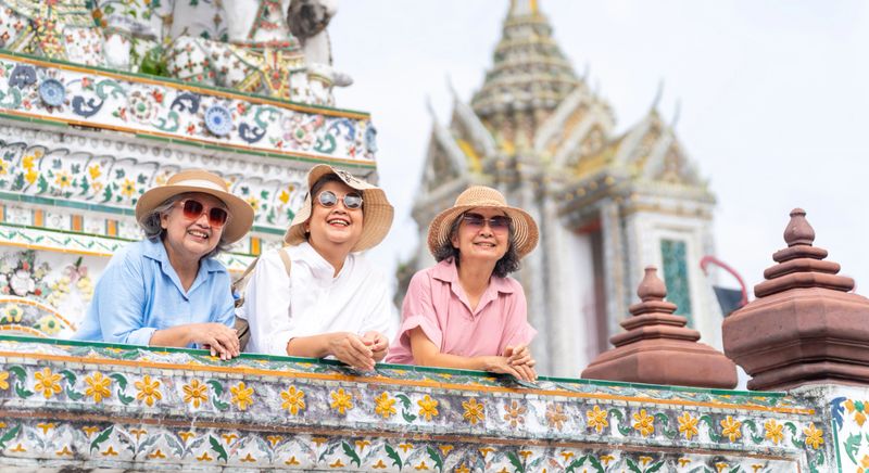 Group of Happy Asian senior woman tourist enjoy and fun outdoor lifestyle travel Bangkok city, Thailand on summer holiday vacation. Elderly retired women friends looking at beautiful architecture of Buddhist temple.