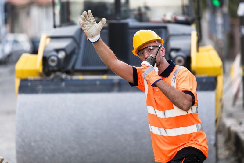 A construction worker in a bright safety vest and hard hat is signaling for vehicles to stop while overseeing roadwork in a busy urban area.