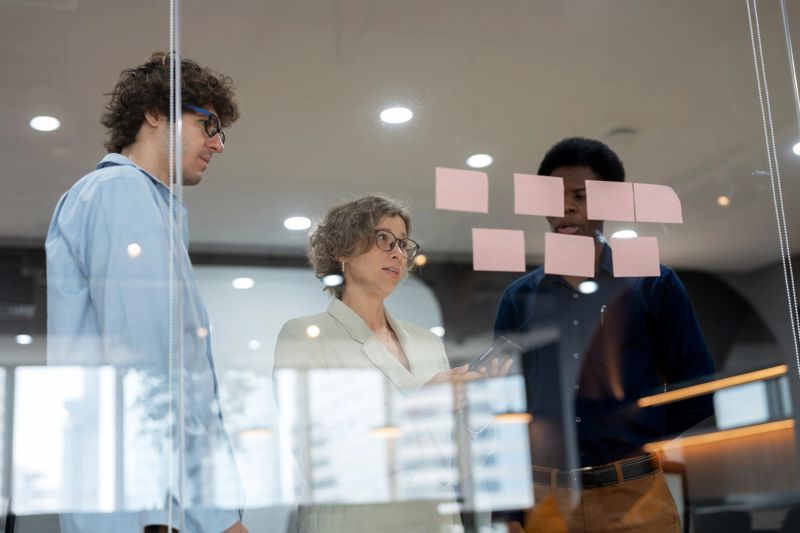Diverse group of business development brainstorming product development ideas in a meeting room, using sticky notes on a glass wall. Collaboration, creativity, and teamwork drive innovation, business strategy, and digital transformation in a modern corporate environment.