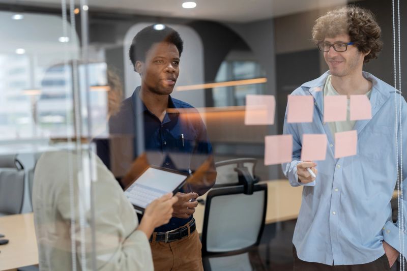 Head of product development standing in a meeting room, discussing ideas with a multiracial team while posting adhesive notes on the glass wall. The session focuses on analyzing market data, creating strategies, and driving business innovation through collaboration and teamwork.