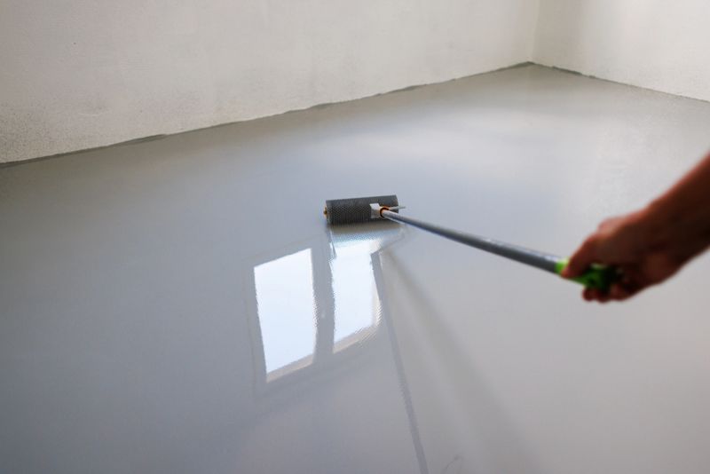 A close-up of a hand using a spiked roller to apply epoxy resin coating on a newly leveled floor. The reflective surface highlights the professional finishing process in a bright, natural-light environment. Ideal for illustrating industrial flooring, home renovation, or construction projects.