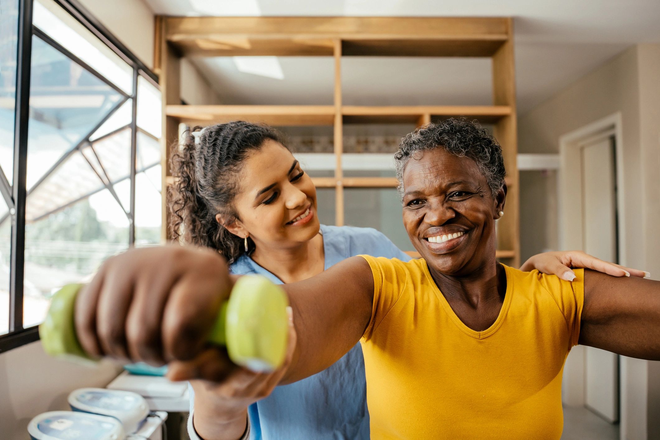 A woman assists an elderly lady with arm exercises using a green dumbbell indoors. Haines City therapy, personalized rehabilitation, physical therapy services