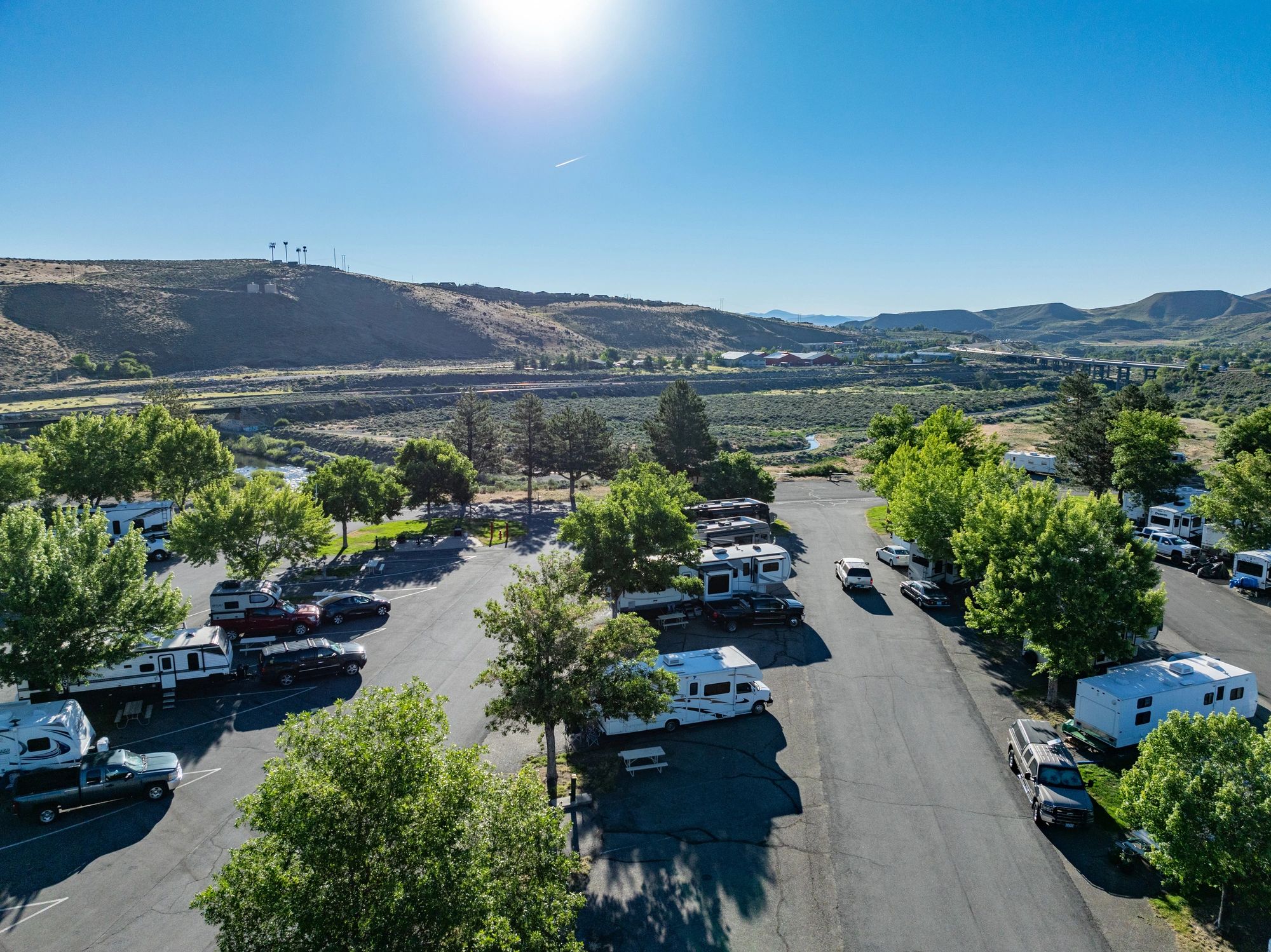 RV park with several campers parked among green trees, set against sunlit hills and a clear blue sky