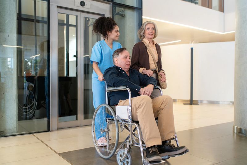 Nurse pushes an elderly man in a wheelchair through a hospital hallway with his wife walking along. Caring nurse with senior patient on wheelchair with wife in hospital corridor.