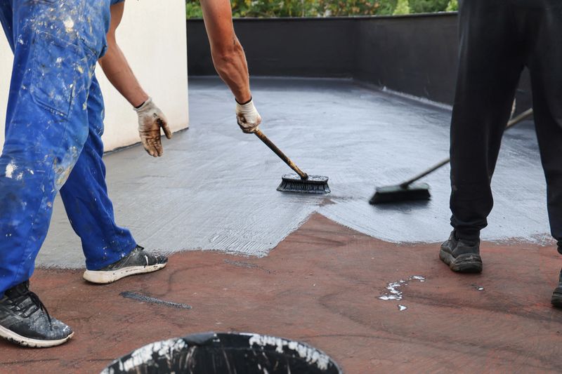 Two builders construction workers with brushes applying waterproofing layer coating on a building roof