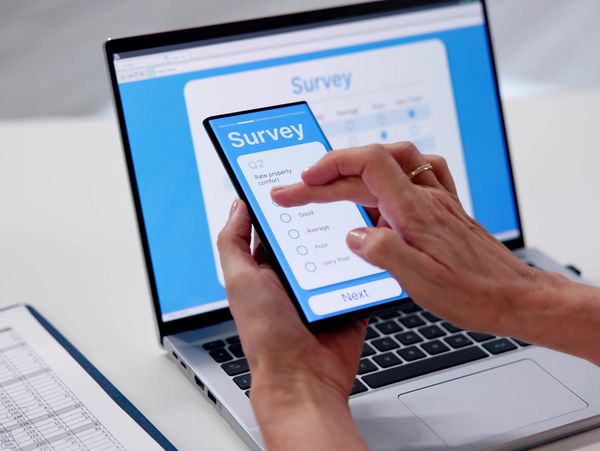Person completing an online survey on a smartphone with a laptop in the background.