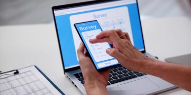 Person completing an online survey on a smartphone with a laptop in the background.