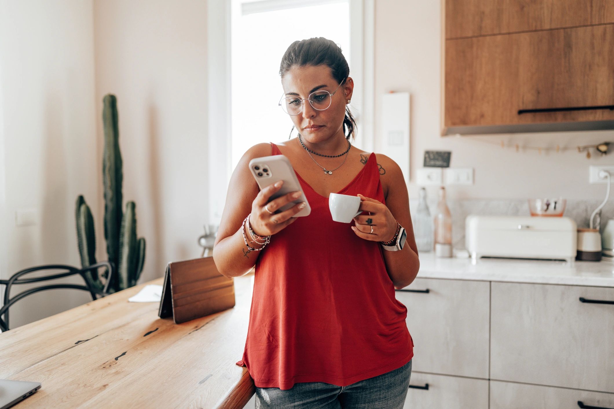 Woman in red top using phone and holding a cup in a cozy kitchen.
