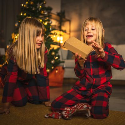 Two children in red plaid pajamas opening Christmas presents by the tree.