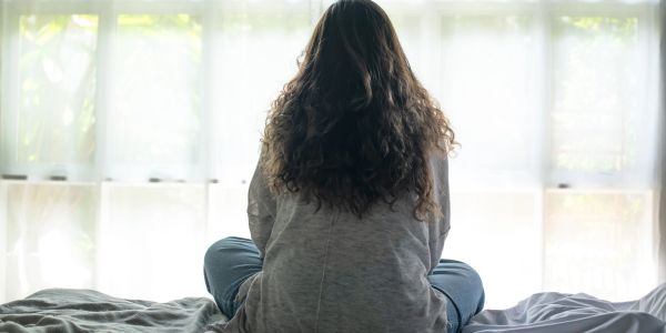 Woman sitting on bed facing a window with natural light.