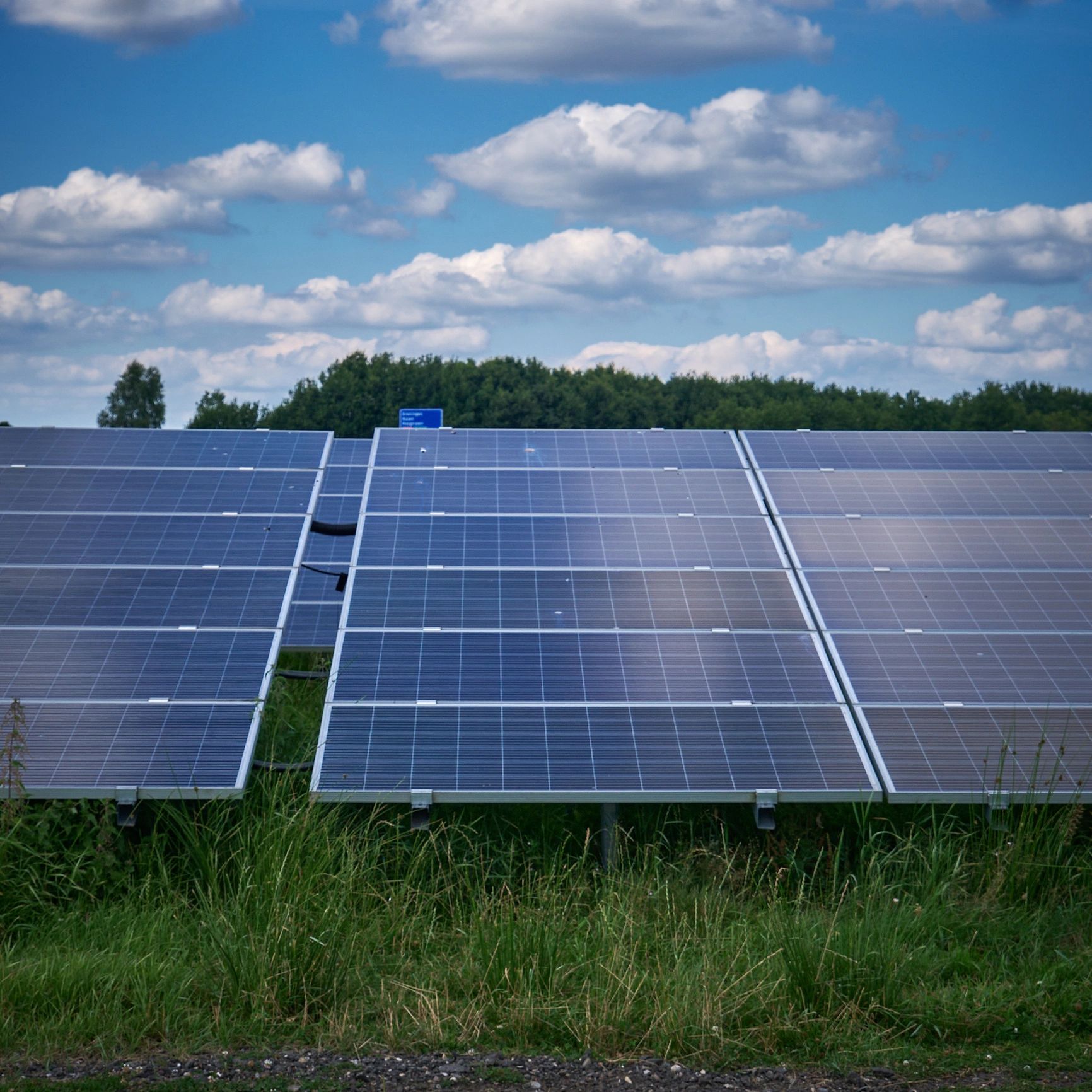 Solar panels under a blue sky with clouds and greenery.