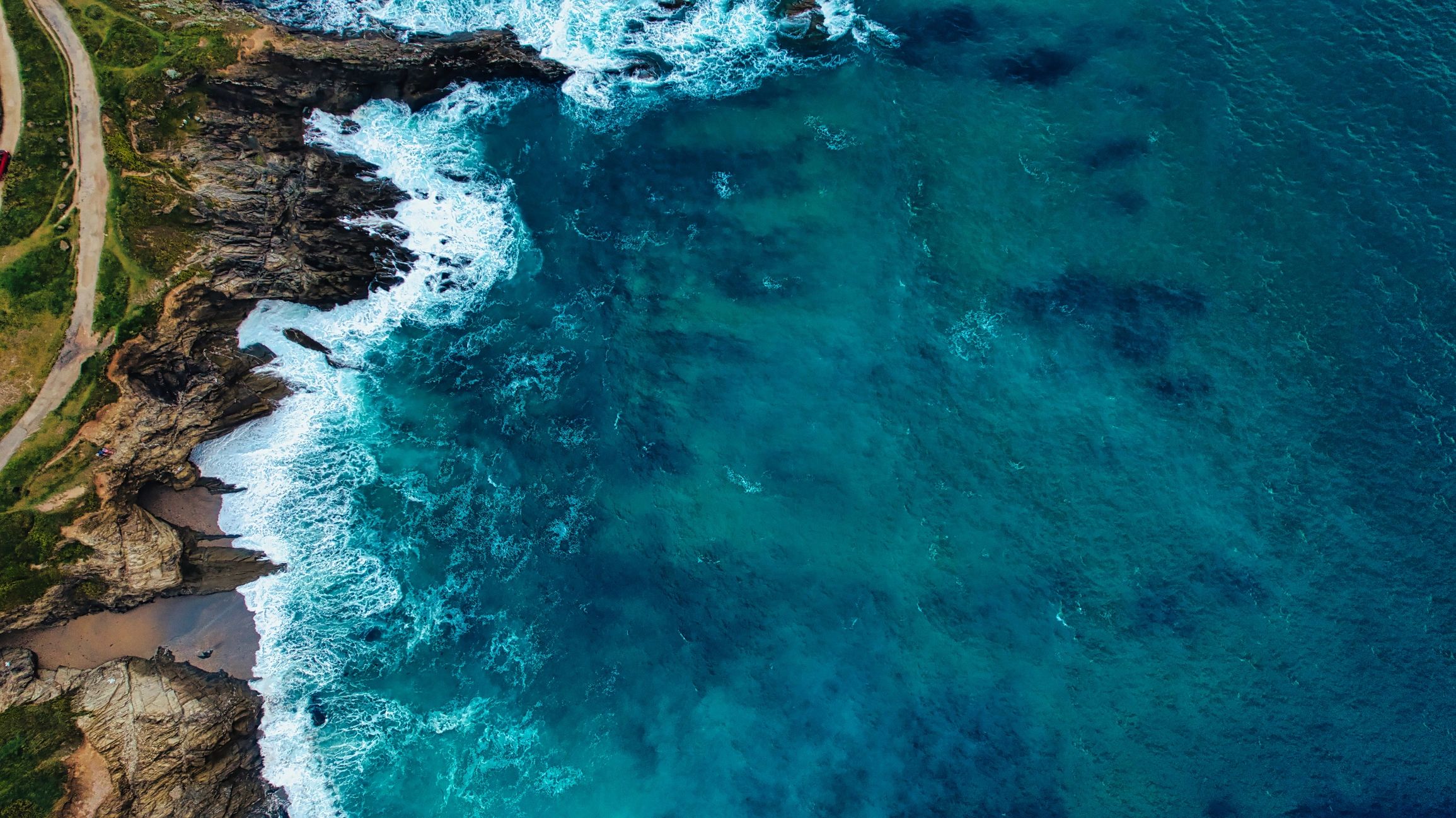 Aerial view of waves crashing against rocky coastline with a dirt path.