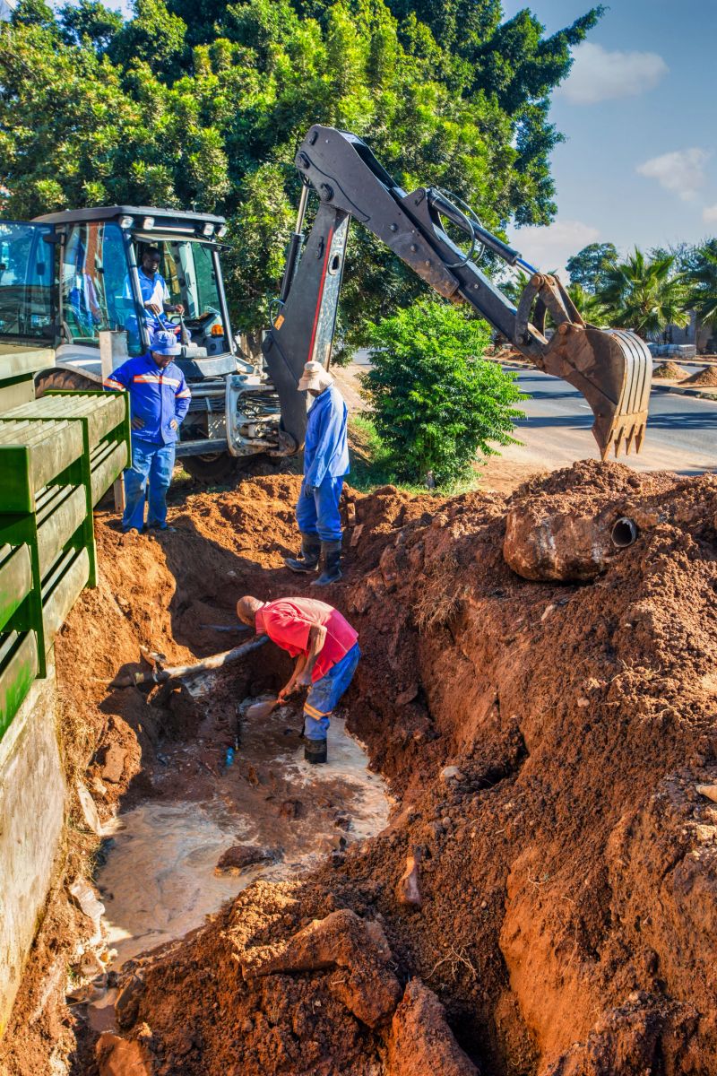 african american construction workers with an excavator fixing a blocked damaged broken drainage pipe in the trench outdoors