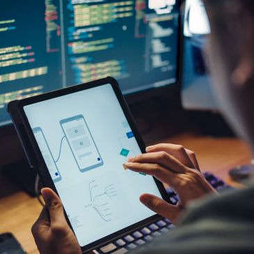 Person working on app design on a tablet with code on a monitor in the background.