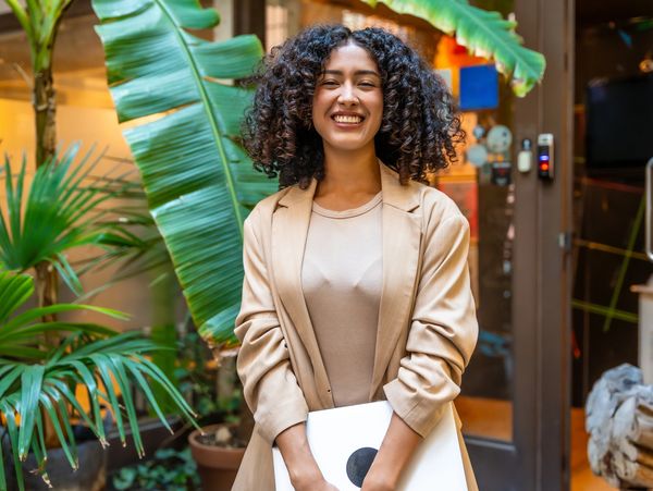 Smiling woman in beige blazer holding a laptop, standing among lush green plants.