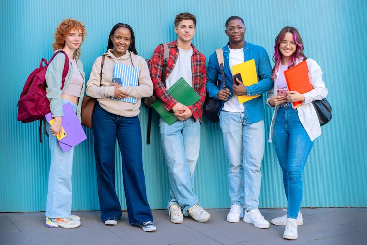 Five diverse students standing against a blue wall, holding folders and smiling.