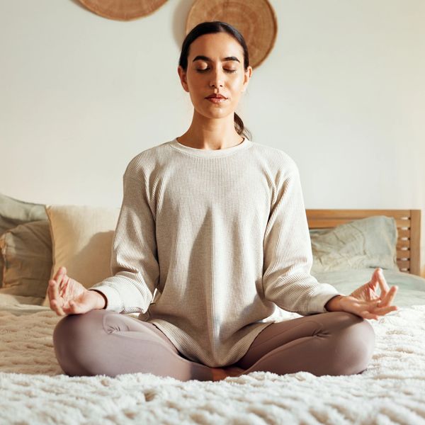 Woman meditating cross-legged on a bed in a calm, cozy room.