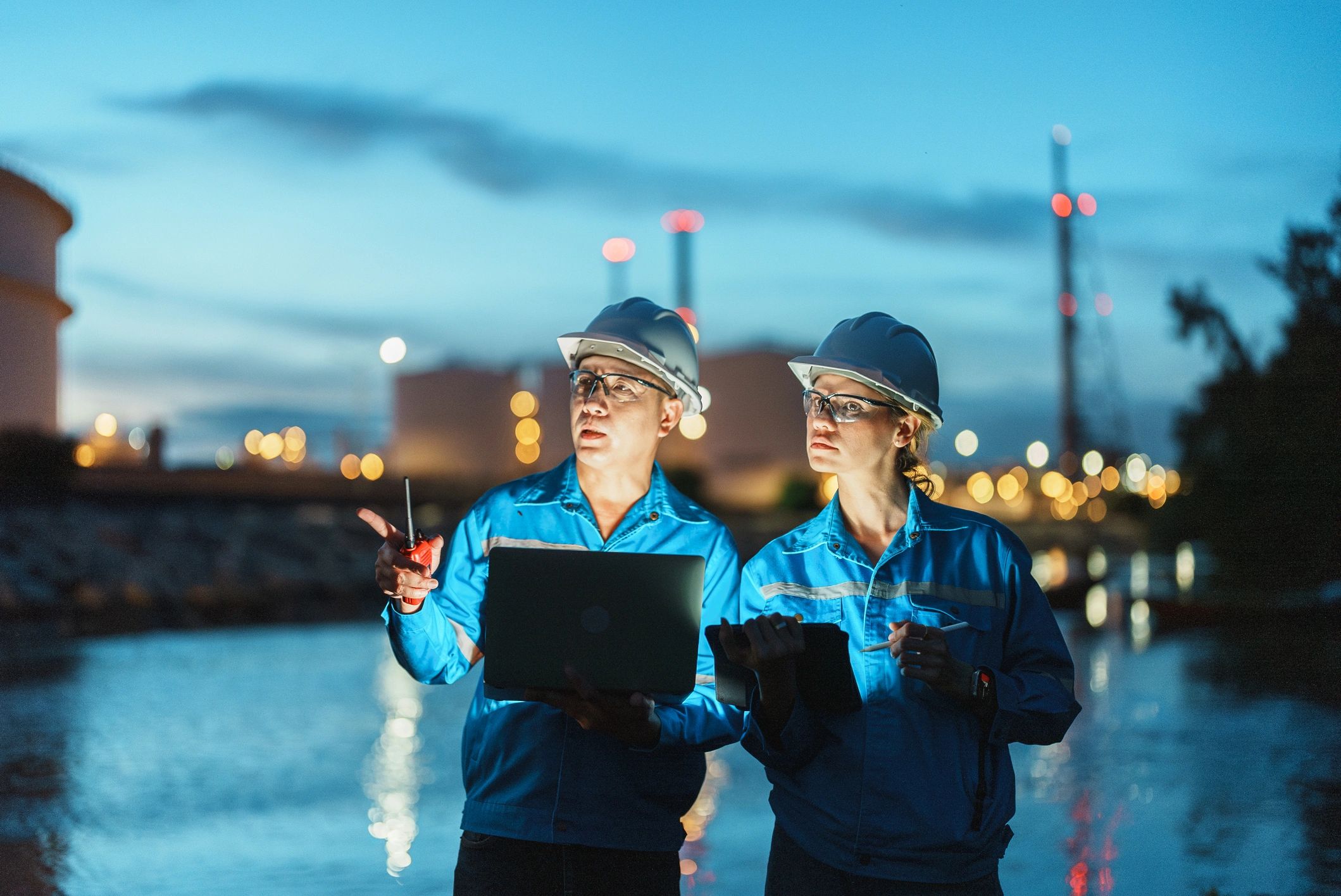 Two engineers in blue uniforms and helmets discussing work at an industrial site at dusk.