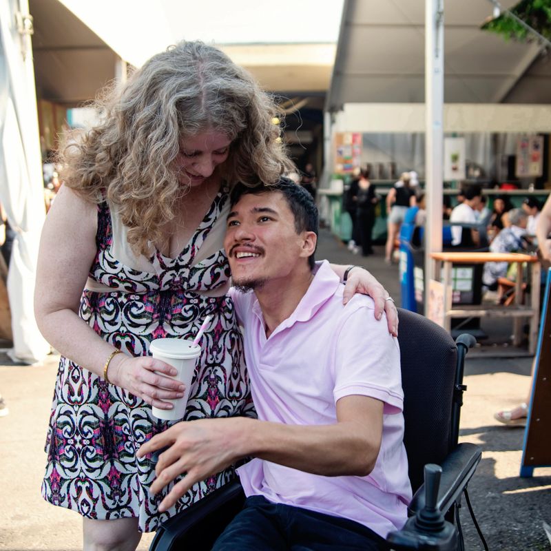 Man living with cerebral palsy with his girlfriend in a public market. He is sitting in his electric wheelchair, and wearing a pink polo shirt. She has a printed pink summer dress, holding his drink. Square waist up outdoors shot. This is part of a series shot in Montreal, Quebec, Canada.