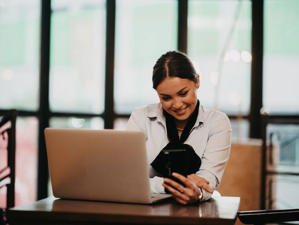 A Latina businesswoman sits in a bustling coffee, skillfully managing communications.