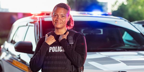 A confident female police officer stands in front of a police car with flashing lights.